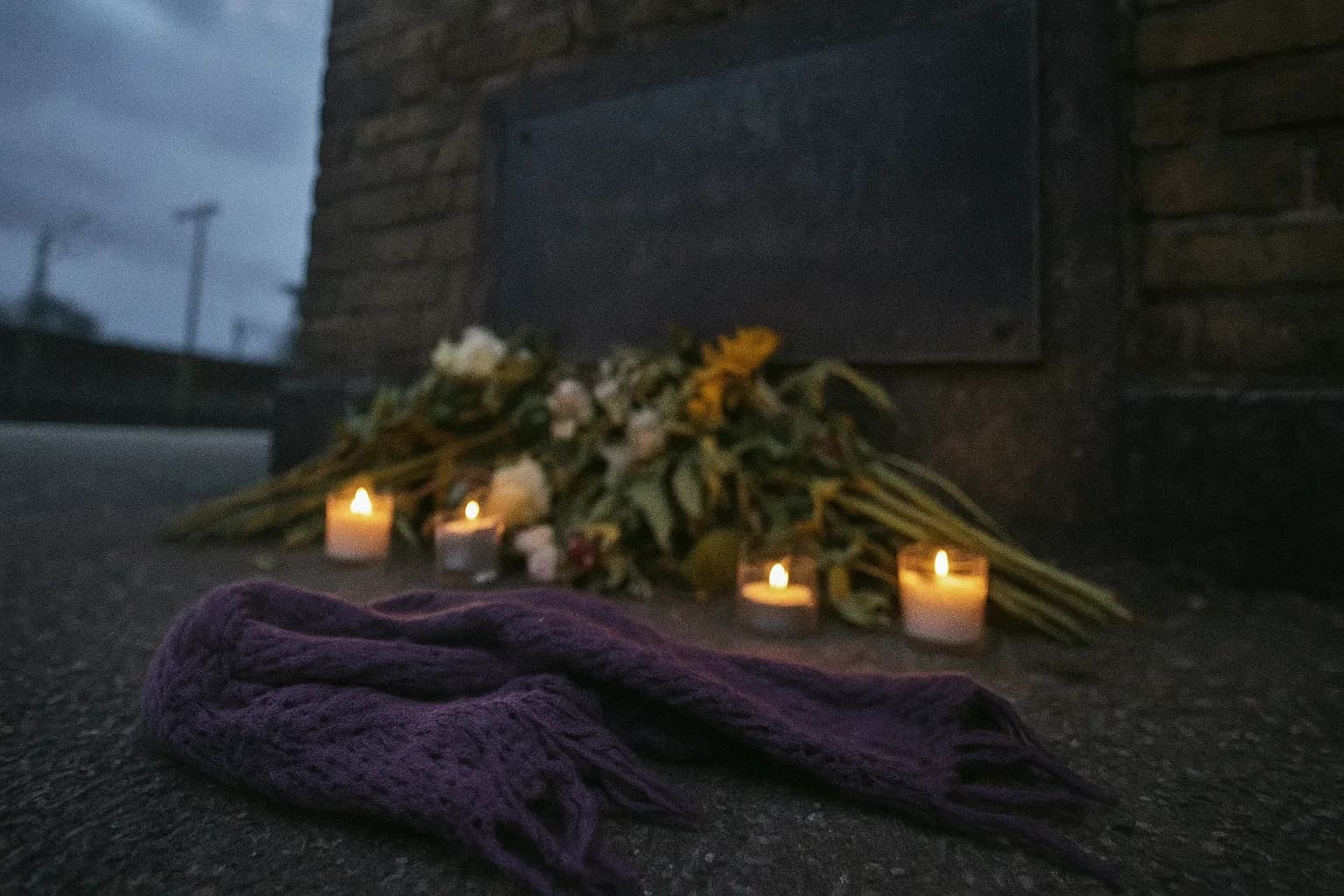 En slidt lilla tørklæde ligger ved foden af en mindeplaque på Korsør Station, omgivet af visne blomster og små lys, der blafrer i vinden.